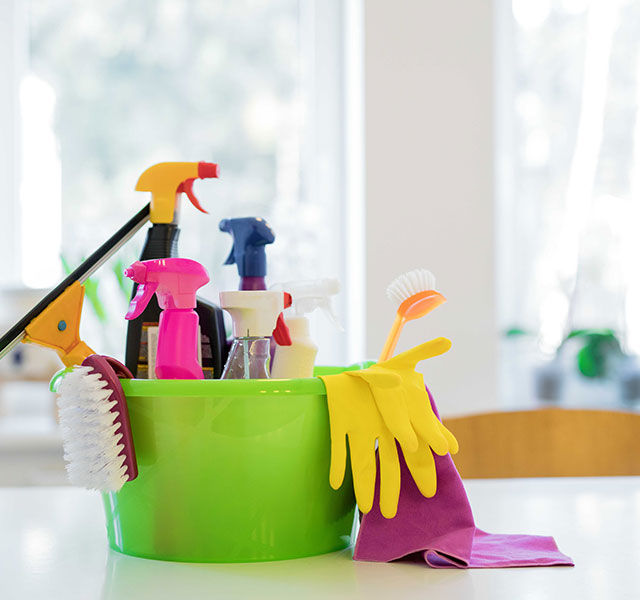 A woman in a white shirt and yellow gloves is cleaning a window with a cloth, while two people in the background are tidying up an office space. One is using a vacuum cleaner, and the other is working at a desk.